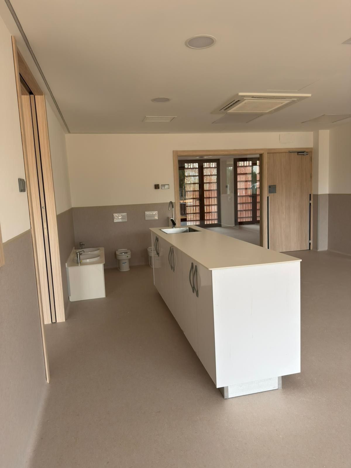 Habitación moderna y vacía con isla blanca, lavabo y acceso a otras habitaciones. Tonos neutros claros.