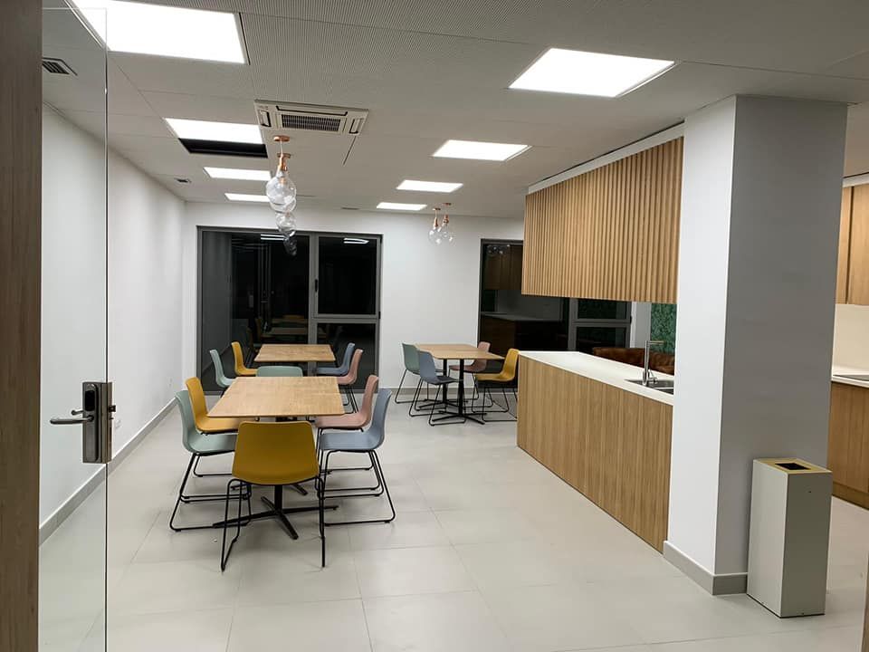 Moderna sala de descanso con mesas, sillas y cocina americana. Tonos neutros y buena iluminación.