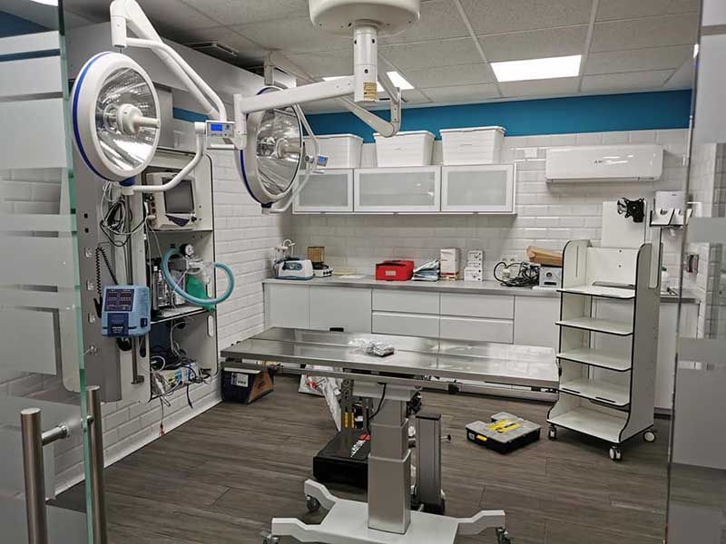 Una sala de operaciones con luces quirúrgicas sobre una mesa de acero inoxidable, gabinetes blancos y equipo médico.