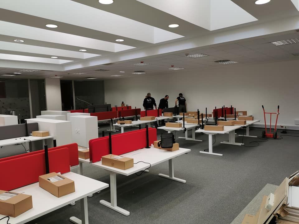 Oficina con filas de escritorios, separadores rojos y cajas de cartón. Tres personas de pie al fondo.