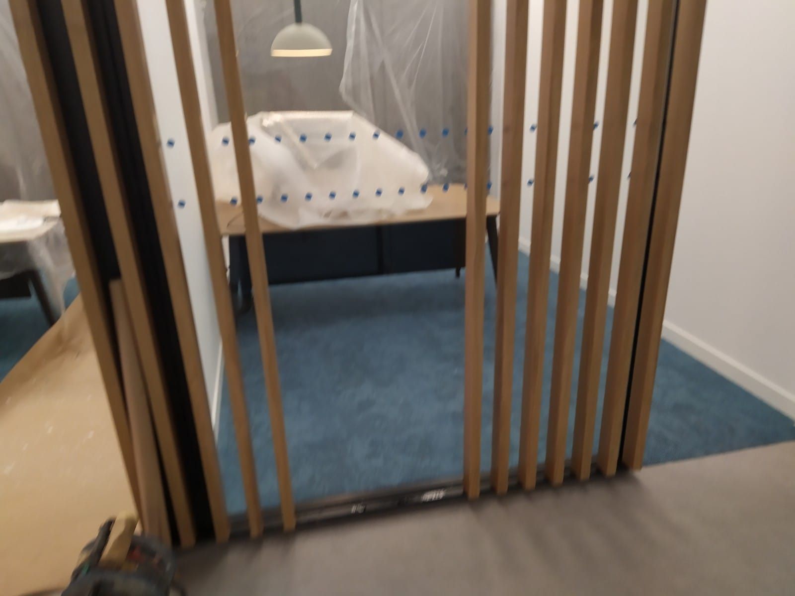 Una mampara de listones de madera oculta parcialmente una habitación con una alfombra azul, un escritorio y un objeto tapado.