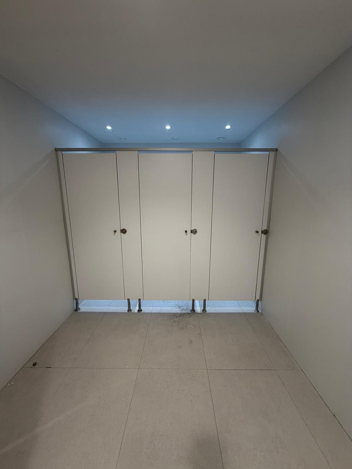 Tres cabinas de baño blancas en un espacio estrecho de tonos neutros; iluminación empotrada.