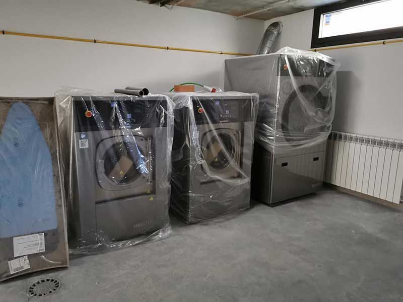 Lavandería con tres lavadoras industriales y una secadora envueltas en plástico. Se ve una tabla de planchar.