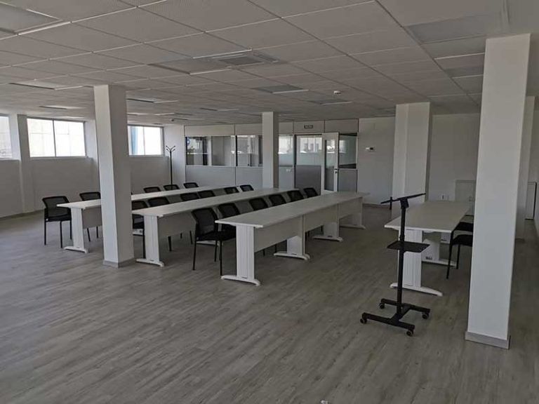 Sala de conferencias vacía con largas mesas, sillas y columnas blancas. Suelo gris y paredes blancas.