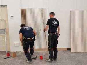 Dos trabajadores instalan paneles verticales en una pared blanca. Uno sostiene una varilla, el otro se arrodilla.