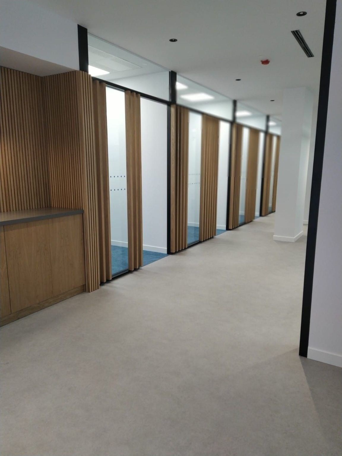 Pasillo con paneles de madera, oficinas con paredes de cristal y paredes blancas. Suelo gris claro.