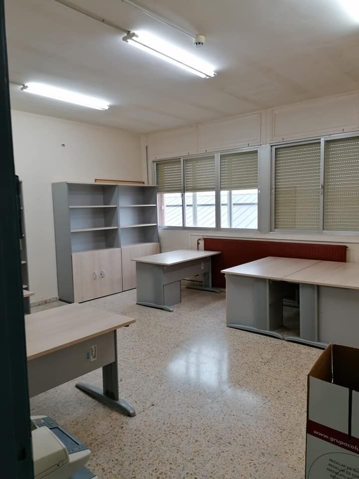 Interior de oficina con escritorios, estanterías y ventanas. Suelo de baldosas. Luces fluorescentes.