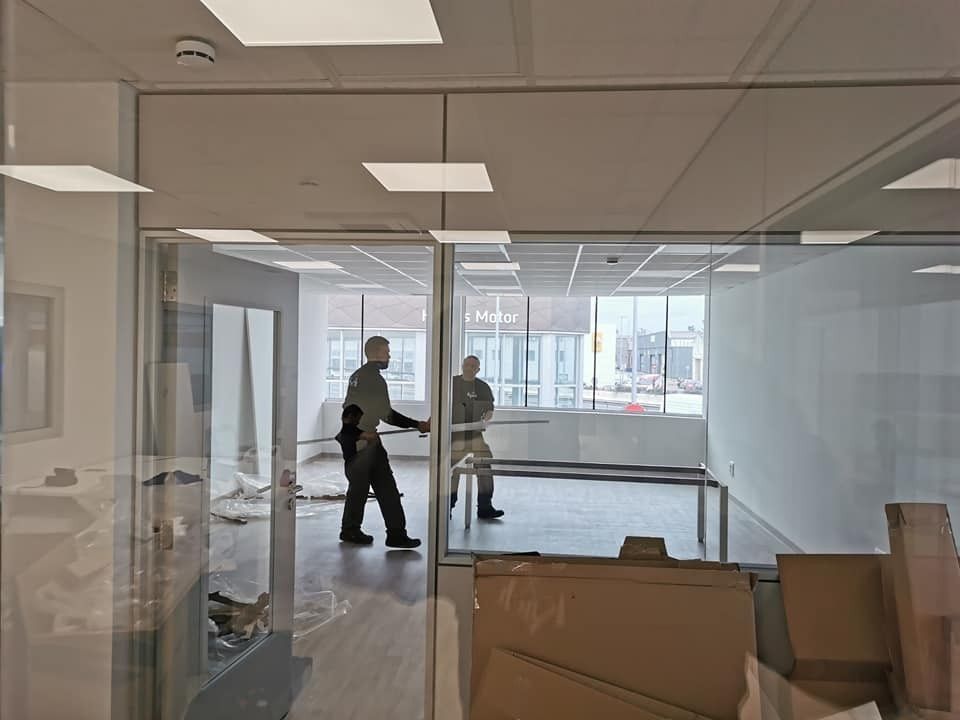 Dos personas en una oficina con paredes de cristal, cajas y una mesa. Una abre una puerta.