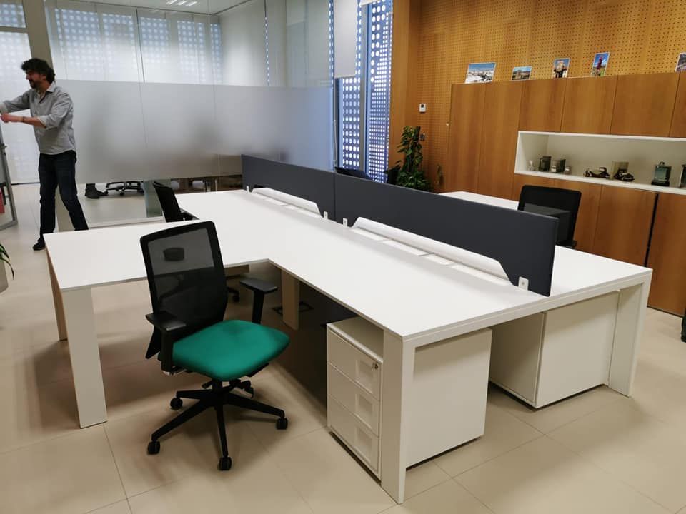 Oficina con escritorios blancos, silla verde azulado y persona organizando objetos; pared y persianas de madera.