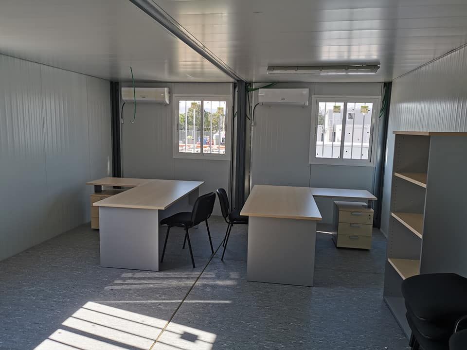 Interior de una oficina modular con escritorios, sillas, ventanas y estanterías. Paredes y suelo grises.