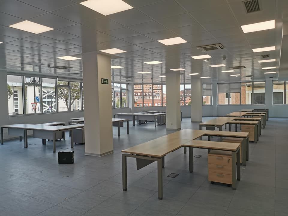 Oficina vacía con filas de escritorios, ventanas e iluminación cenital.