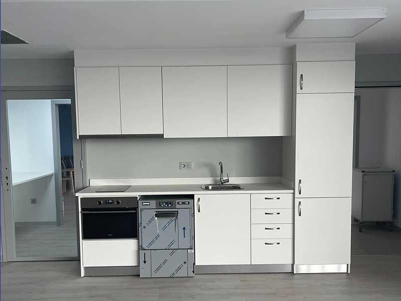 Gabinetes de cocina y electrodomésticos blancos en una habitación con una pared gris claro y un piso de color madera clara.