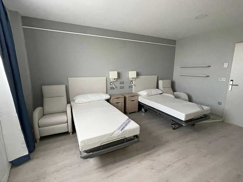 Dos camas de hospital en una habitación de paredes grises, dos mesas de noche