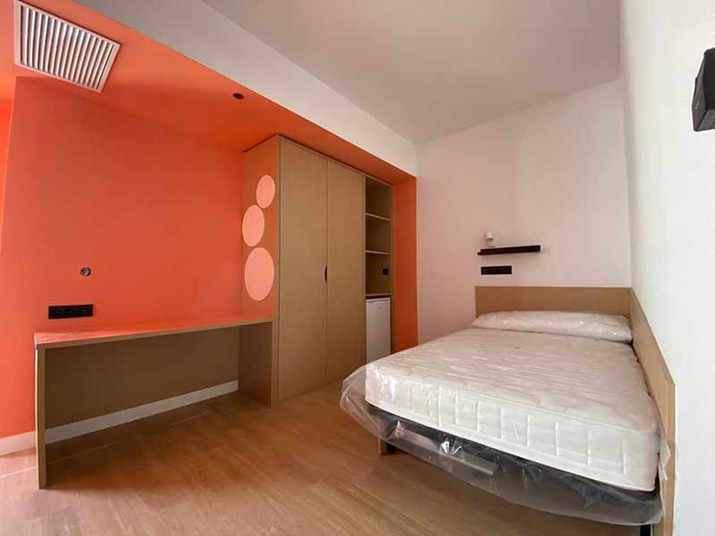 Dormitorio con escritorio, cama, armario y pared con acento naranja. Suelo de madera clara y paredes en tonos neutros.