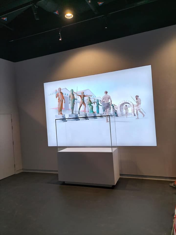 Pantalla iluminada en blanco que muestra figuras con un pedestal blanco en una habitación gris.