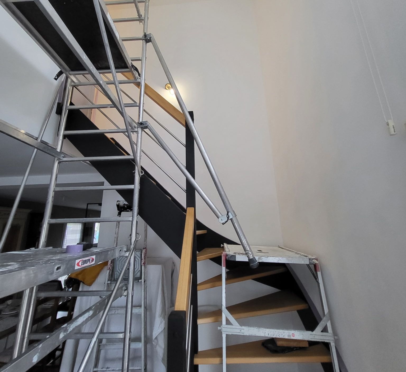 Échafaudage le long d'un escalier, probablement pour des travaux de peinture, dans une pièce avec un mur.