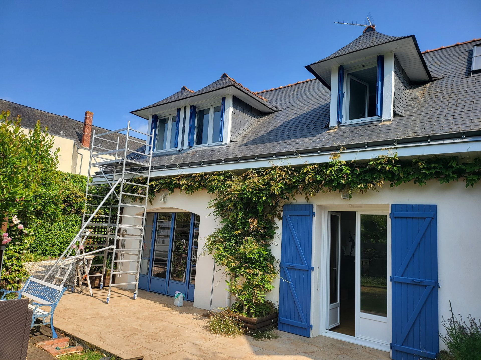 Maison aux volets, deux lucarnes et échafaudages. Du lierre grimpe sur la façade.