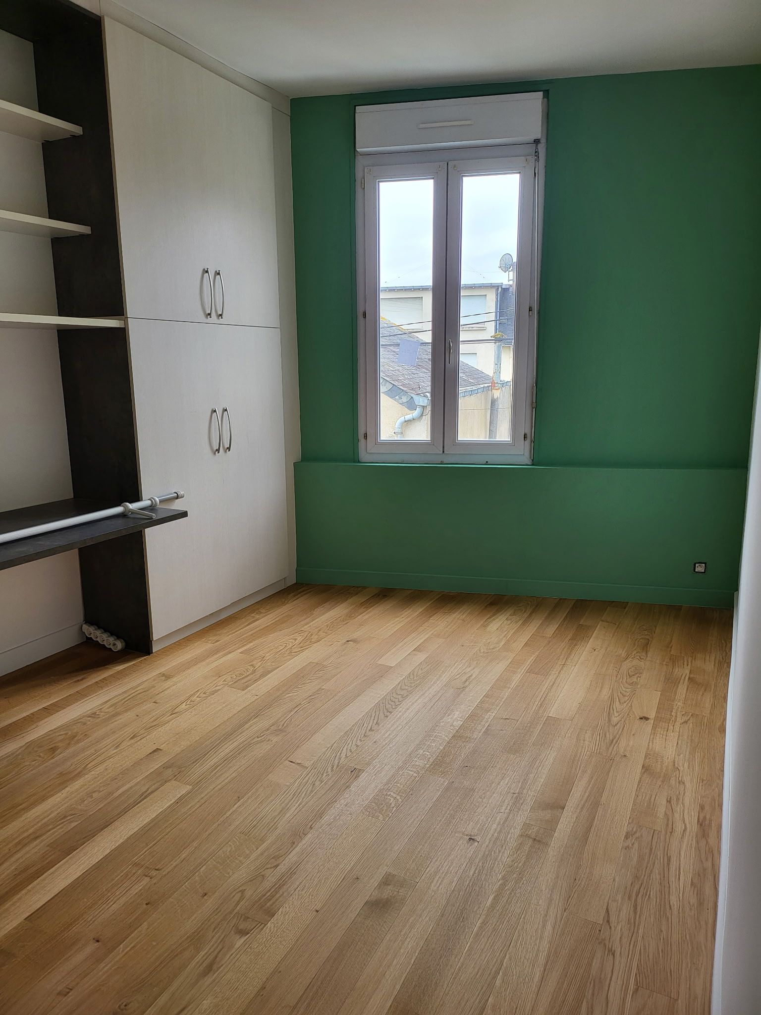 Une pièce lumineuse avec un parquet clair, des étagères et des placards intégrés sur la gauche, et un mur avec une fenêtre.