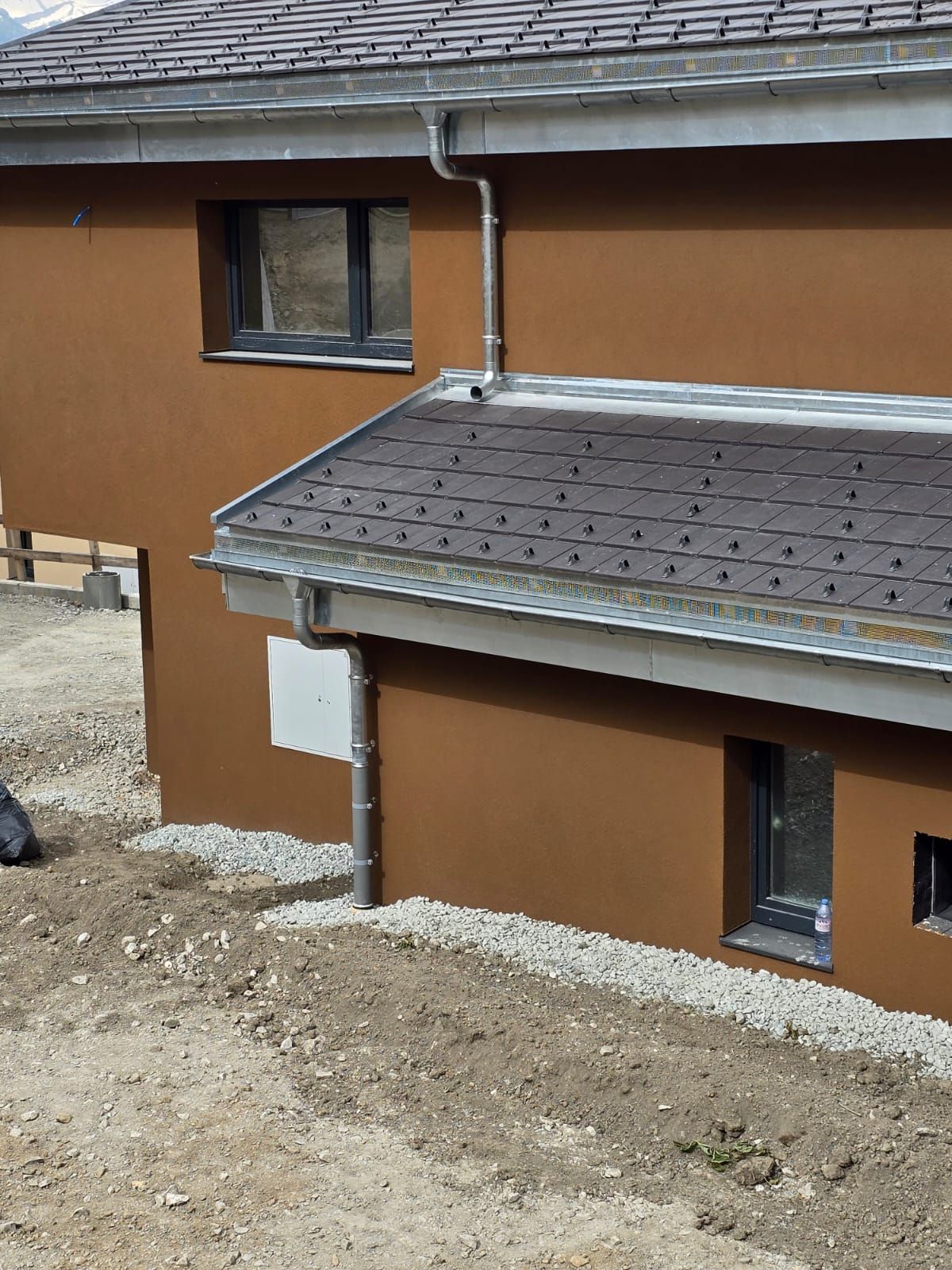 Maison en stuc brun avec toit, fenêtres et gouttières gris ; terrain en gravier.