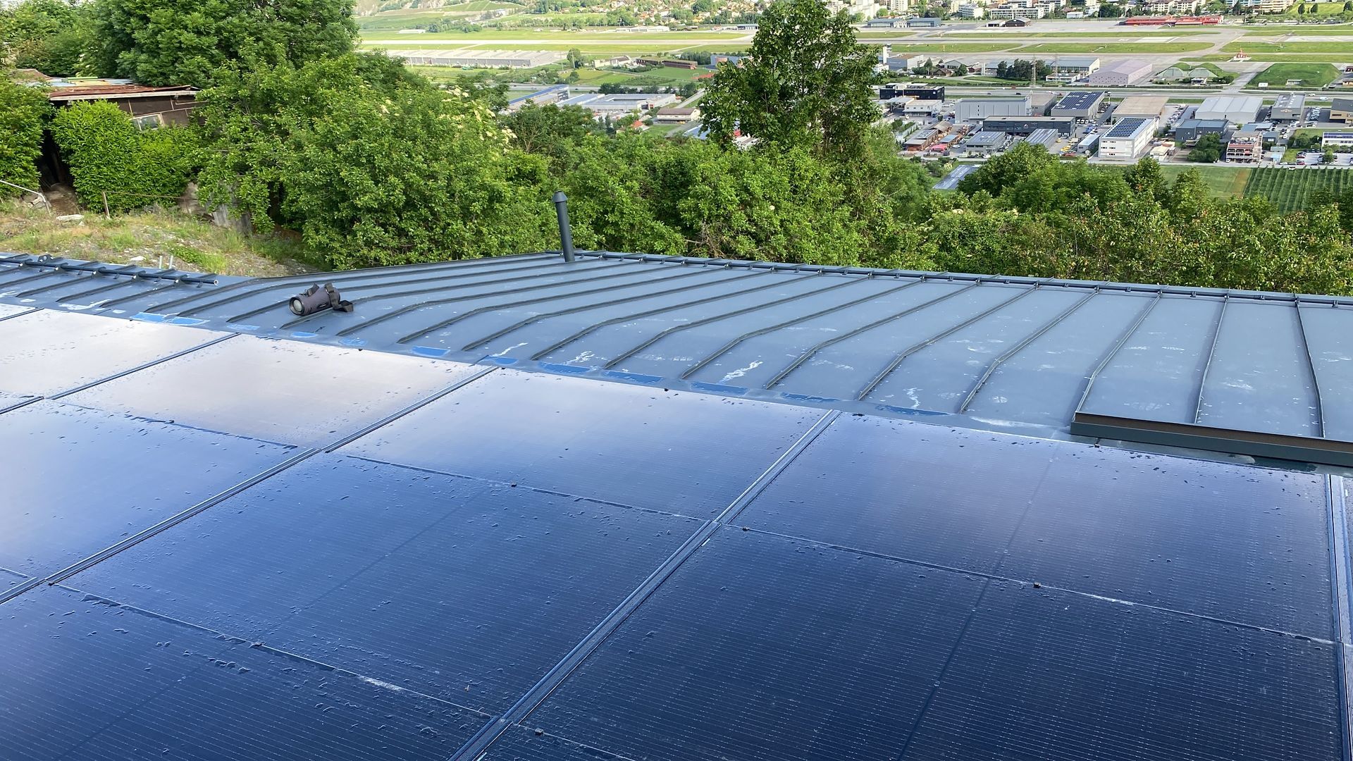 Des panneaux solaires sur un toit en pente reflètent le ciel, avec des arbres et des bâtiments en arrière-plan.
