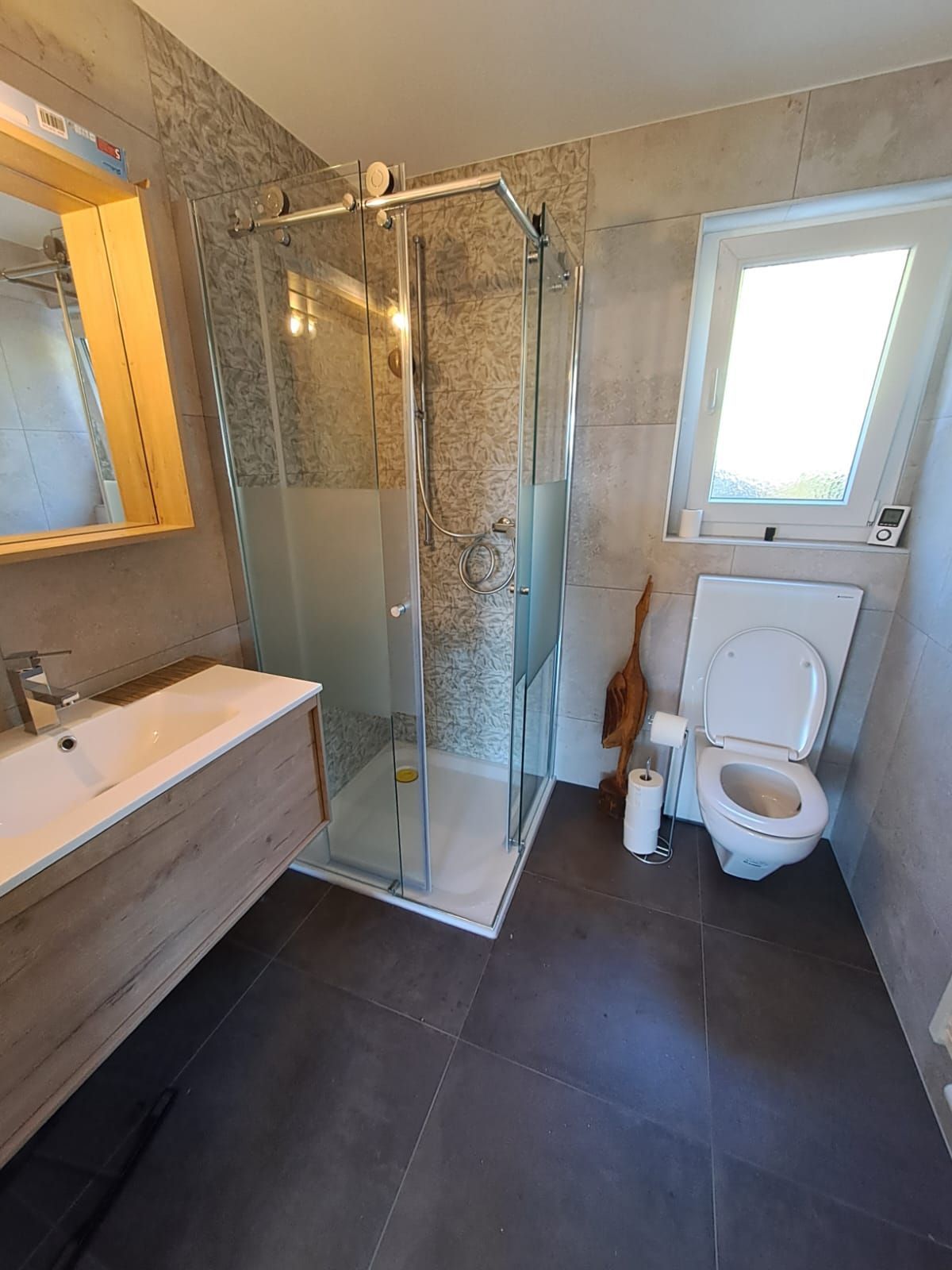 Salle de bains avec toilettes, douche, meuble-lavabo et fenêtre. Carrelage gris et éléments en bois.