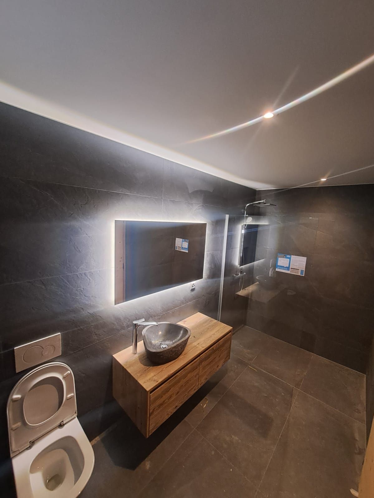 Salle de bains moderne avec murs noirs, meuble-lavabo en bois et miroir rétroéclairé. Toilettes visibles.