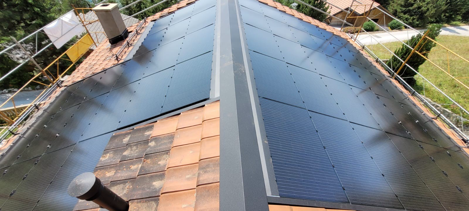 Panneaux solaires sur un toit en tuiles de terre cuite avec une bande centrale. Une cheminée et des gouttières sont également visibles.