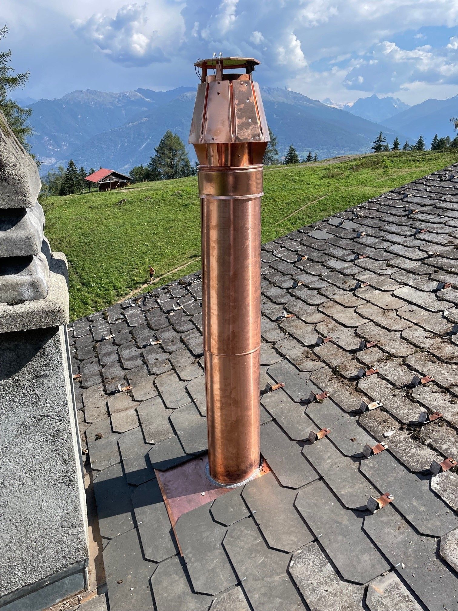 Cheminée en cuivre sur un toit de tuiles, donnant sur une colline verdoyante et des montagnes.