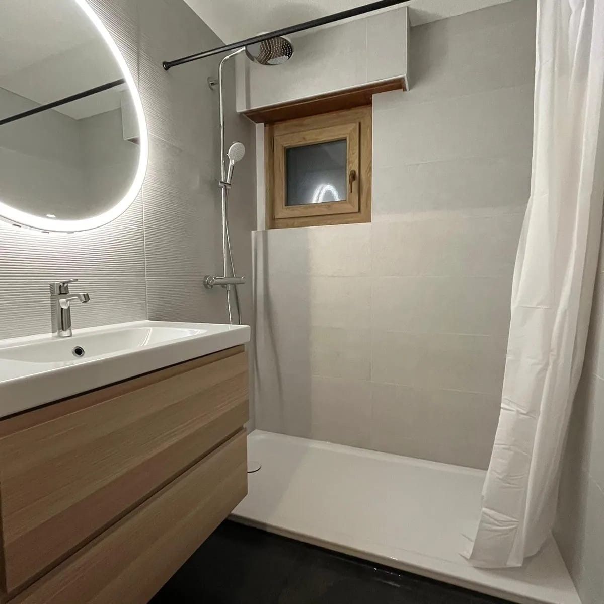 Salle de bains avec meuble-lavabo en bois clair, miroir rond, douche, petite fenêtre et murs gris.