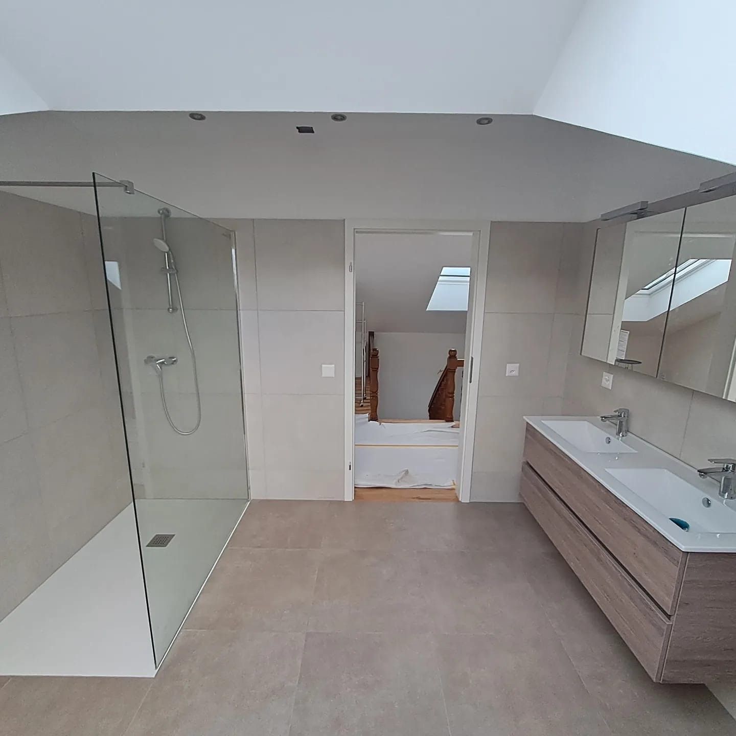 Salle de bains moderne avec douche à l'italienne, double vasque et porte ouverte sur une autre pièce.