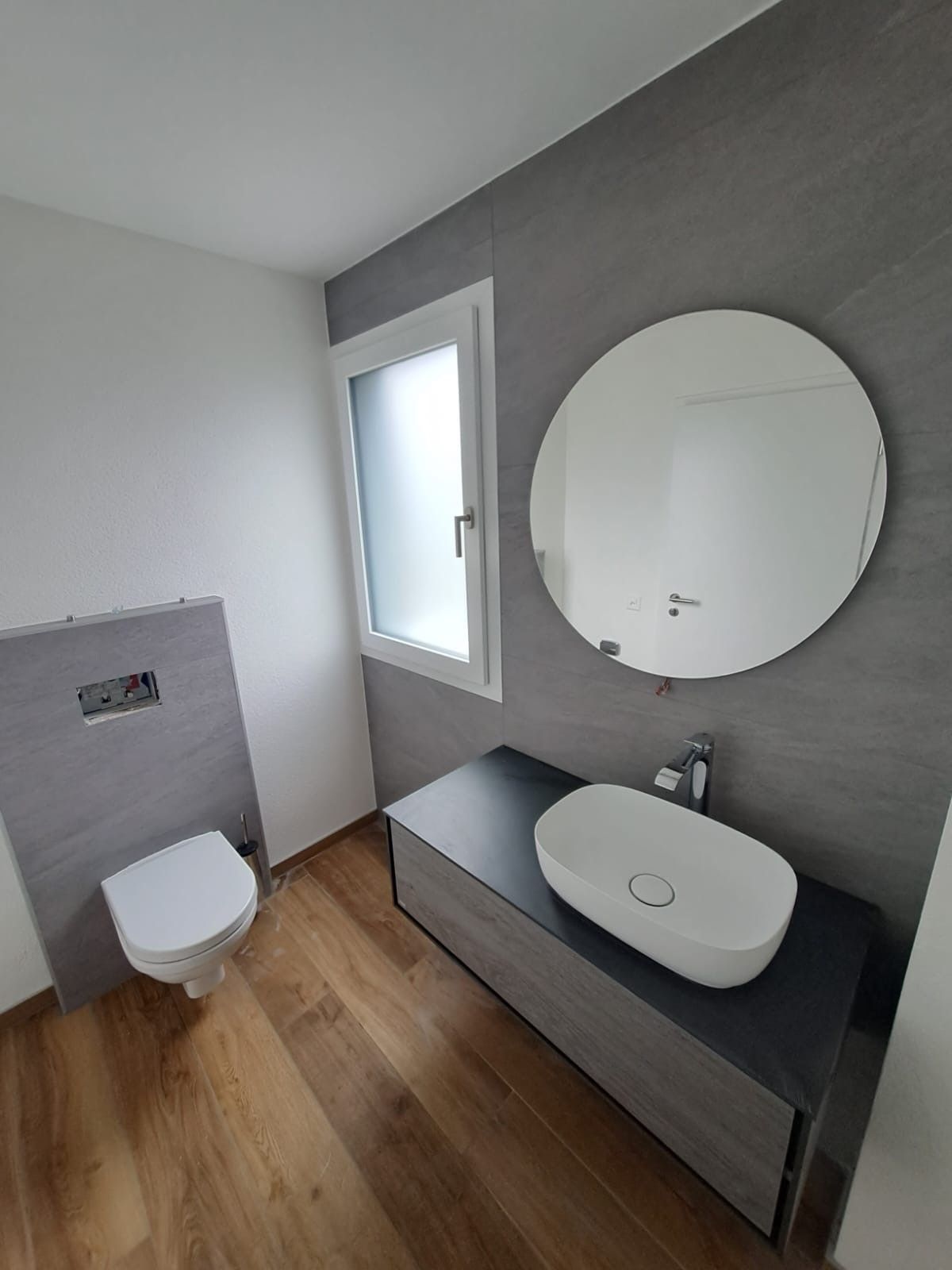 Salle de bains avec meuble-lavabo suspendu, miroir rond, toilettes et fenêtre ; tons gris et bois.