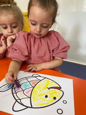 Dos niñas están sentadas en una mesa dibujando un pez con crayones.