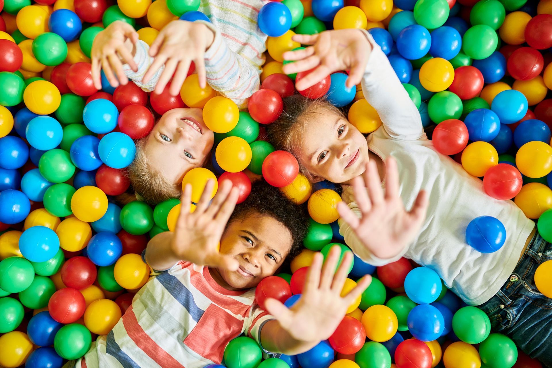 Eine Gruppe von Kindern in bunter Kleidung lächelt und hebt die Arme auf einem sonnigen Spielplatz.