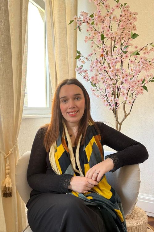 Una mujer con una camisa blanca está sentada frente a una planta en maceta con flores blancas.