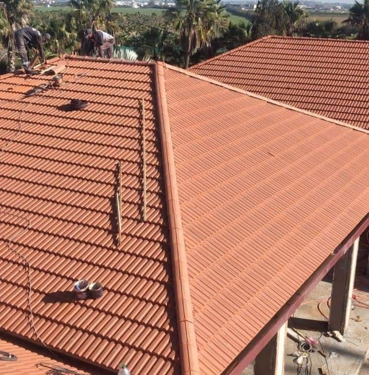 Tejados de tejas rojas en construcción con trabajadores en un tejado inclinado en un barrio soleado.