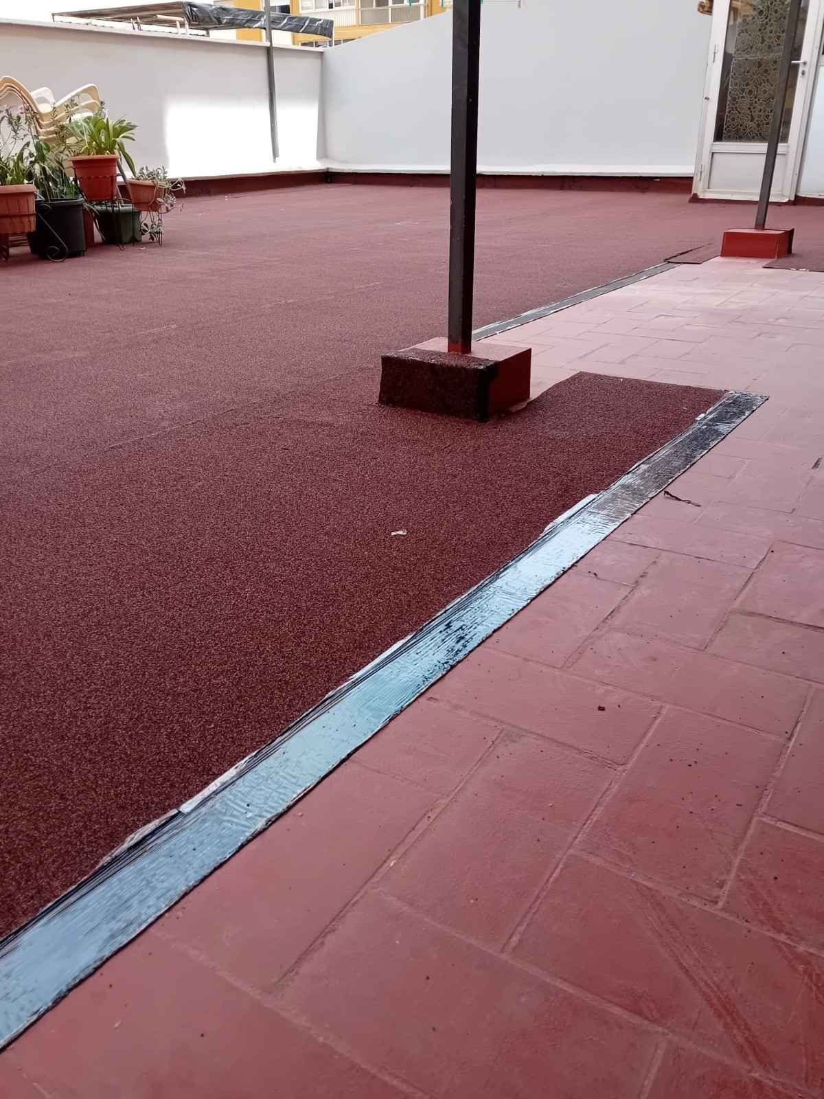Terraza con baldosas rojas, una zona alfombrada de color burdeos, un poste de metal y macetas con plantas a lo largo del borde.