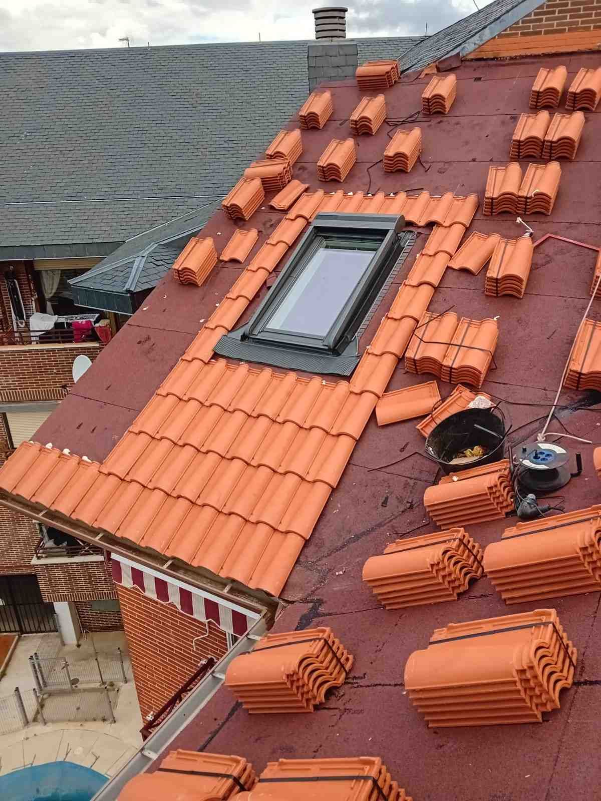 Tejado de tejas rojas con claraboya, chimenea y pilas de tejas de terracota en reparación.