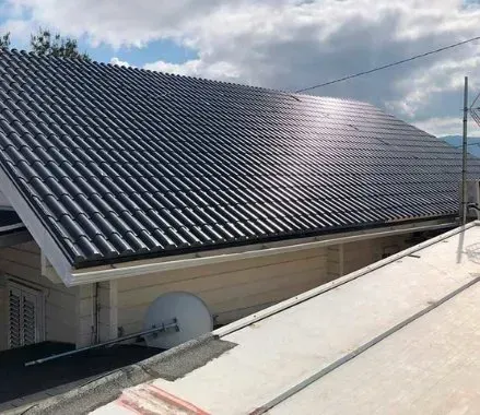 Techo de tejas negras con reflejo del sol sobre una casa beige y una antena parabólica bajo un cielo nublado.