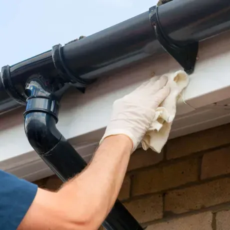 Una mano enguantada limpia con un trapo una canaleta negra en el alero de una casa.