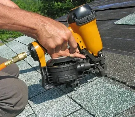 Una mano utiliza una pistola neumática de clavos amarilla para fijar tejas grises del tejado.