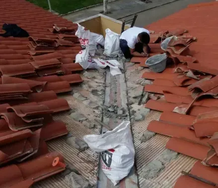 Obreros reemplazando tejas rojas en un tejado dañado, con sacos de construcción y herramientas esparcidas por el suelo.