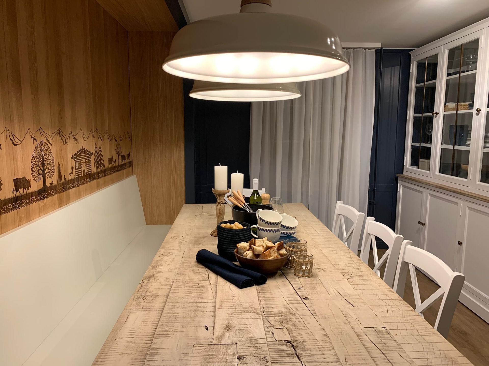 Ein rustikales Esszimmer mit einem langen, gedeckten Holztisch, weißen Stühlen und einer eingebauten Bank. Über dem Tisch hän