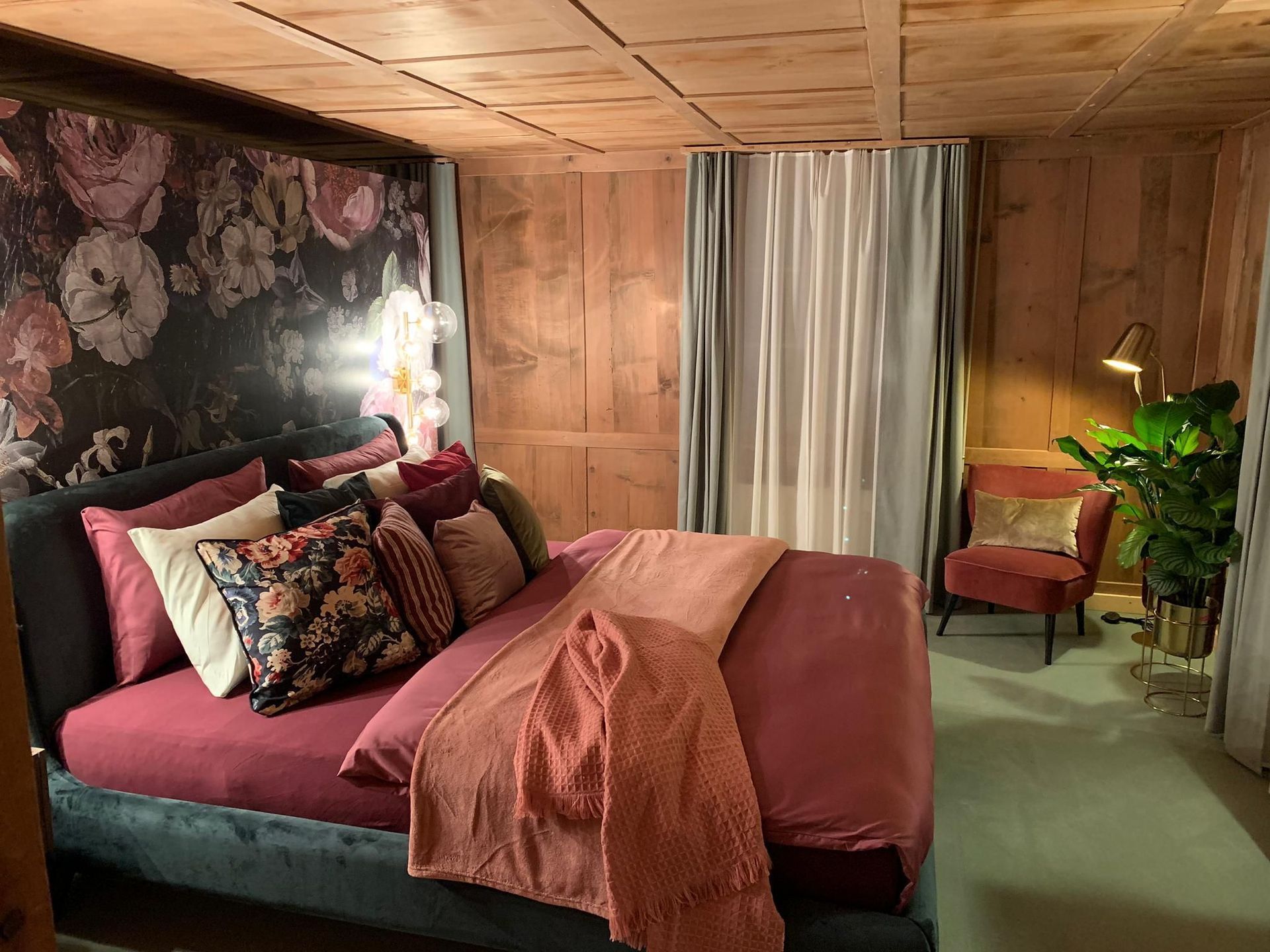 Schlafzimmer mit Samtbett, Blumentapete, Holzvertäfelung und einem Lesesessel mit Stehlampe. Die Farbgebung besteht hauptsäch