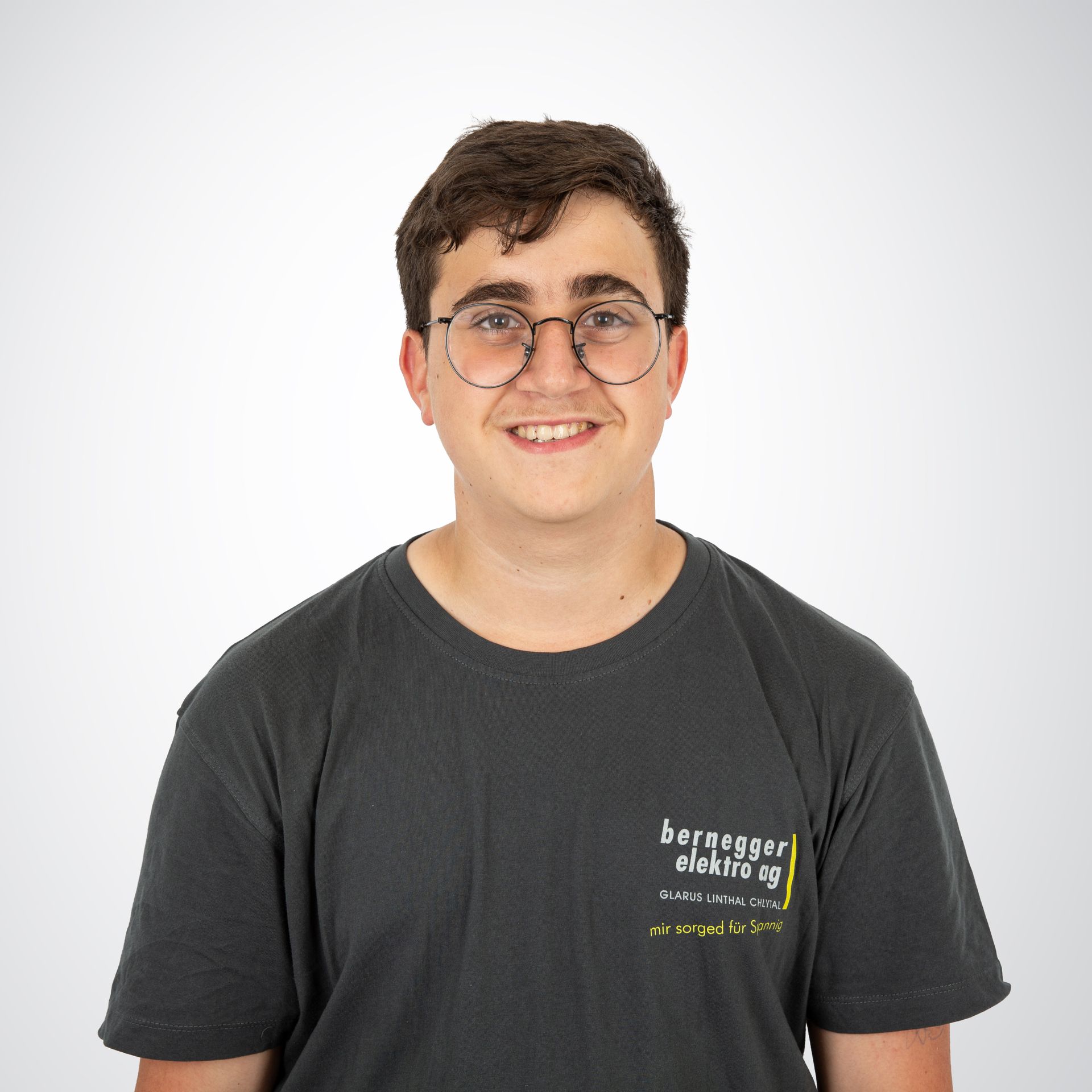 Ein junger Mann mit dunklem Haar und einem freundlichen Lächeln trägt ein graues T-Shirt mit einem Firmenlogo. Auf seinem lin