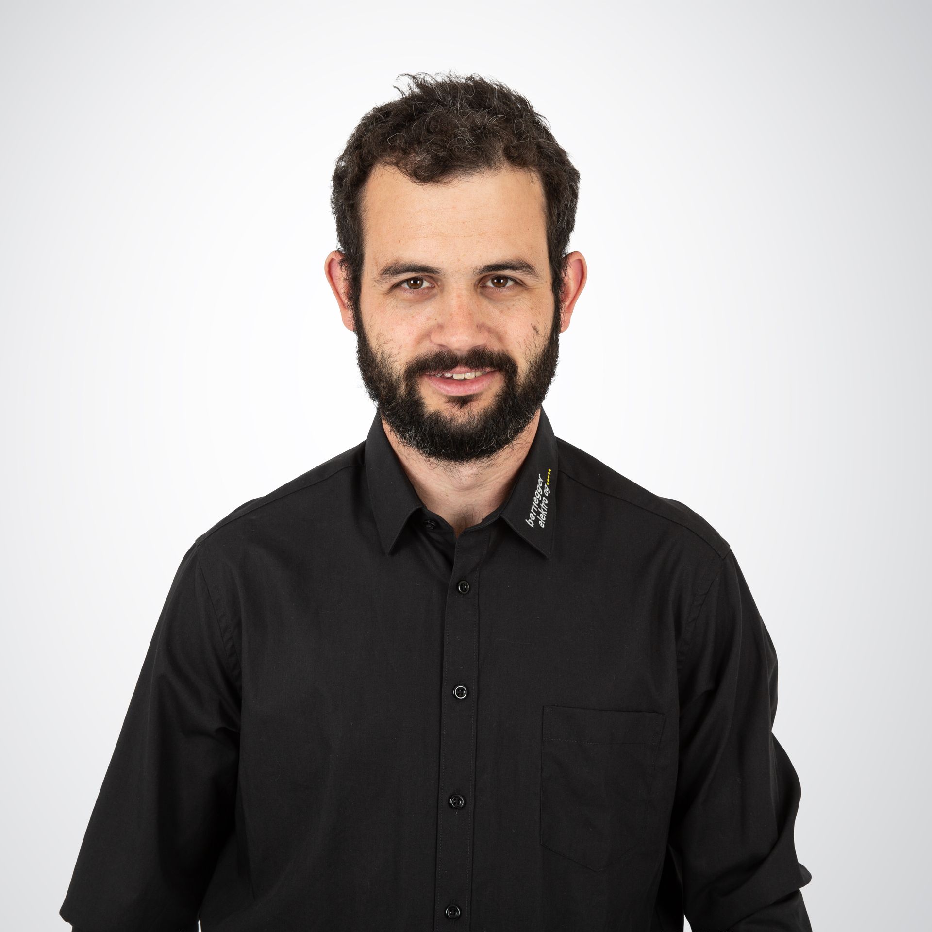 Mann mit Bart, der ein schwarzes Button-Down-Hemd trägt und vor einem weißen Hintergrund in die Kamera lächelt.