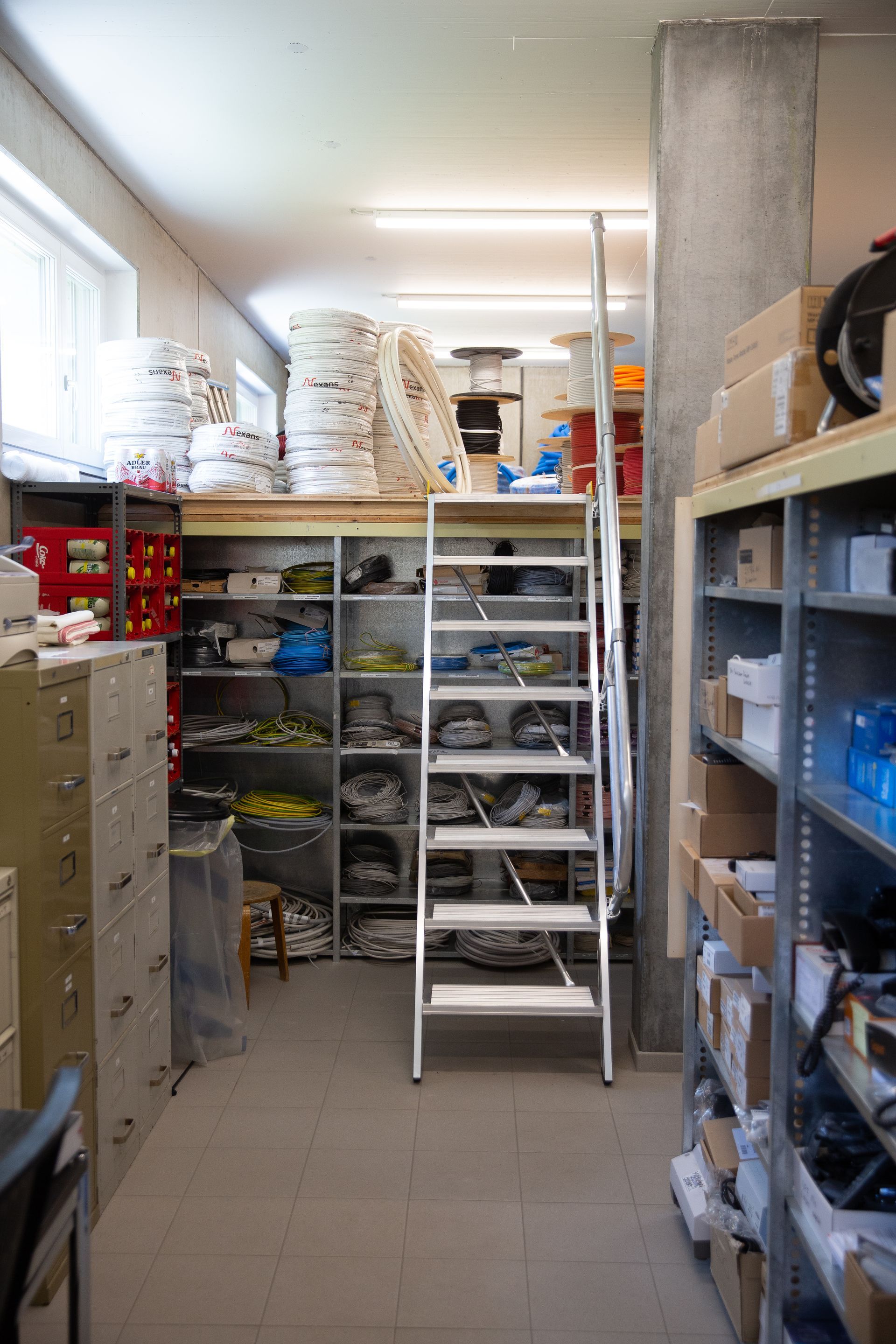 Ein Lagerraum mit Regalen, Aktenschränken und einer Leiter, die zu einem Dachboden führt. Im gesamten Raum werde