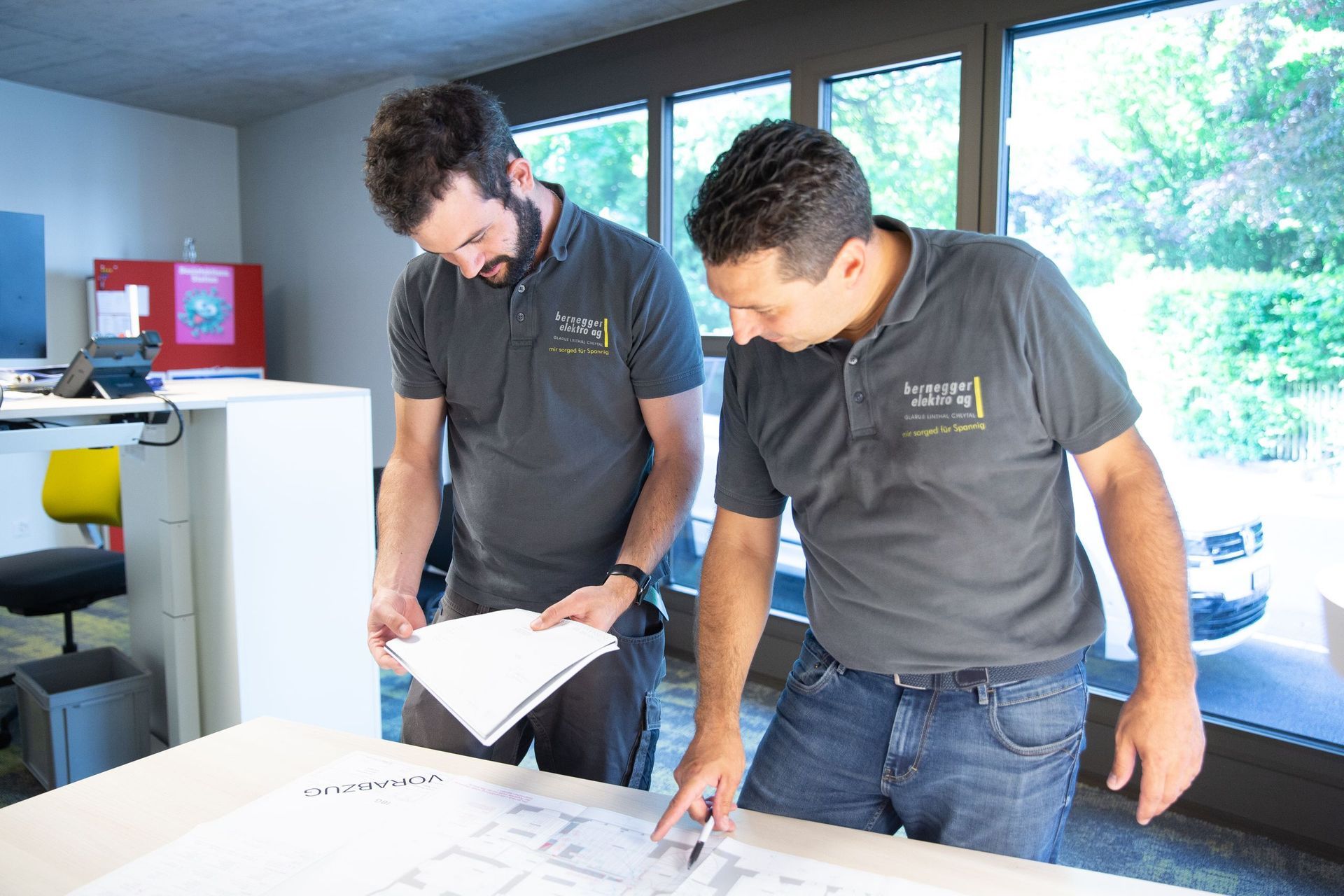 Zwei Männer in grauen Hemden mit Logos betrachten an einem Tisch in einem gut beleuchteten Büro Baupläne.