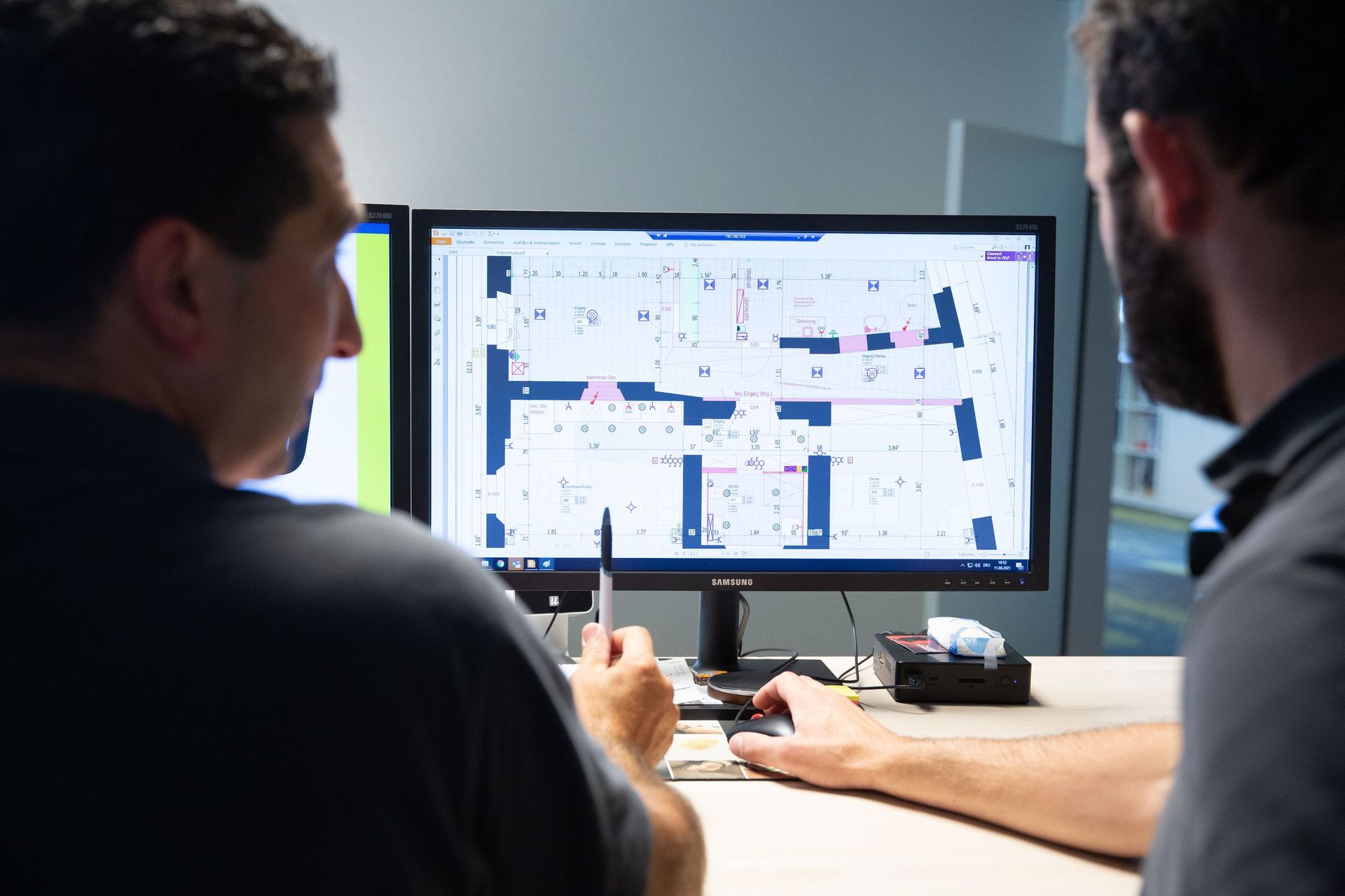 Zwei Männer schauen auf einen Computerbildschirm, auf dem eine Karte angezeigt wird. Einer hält einen Stift, der andere benu