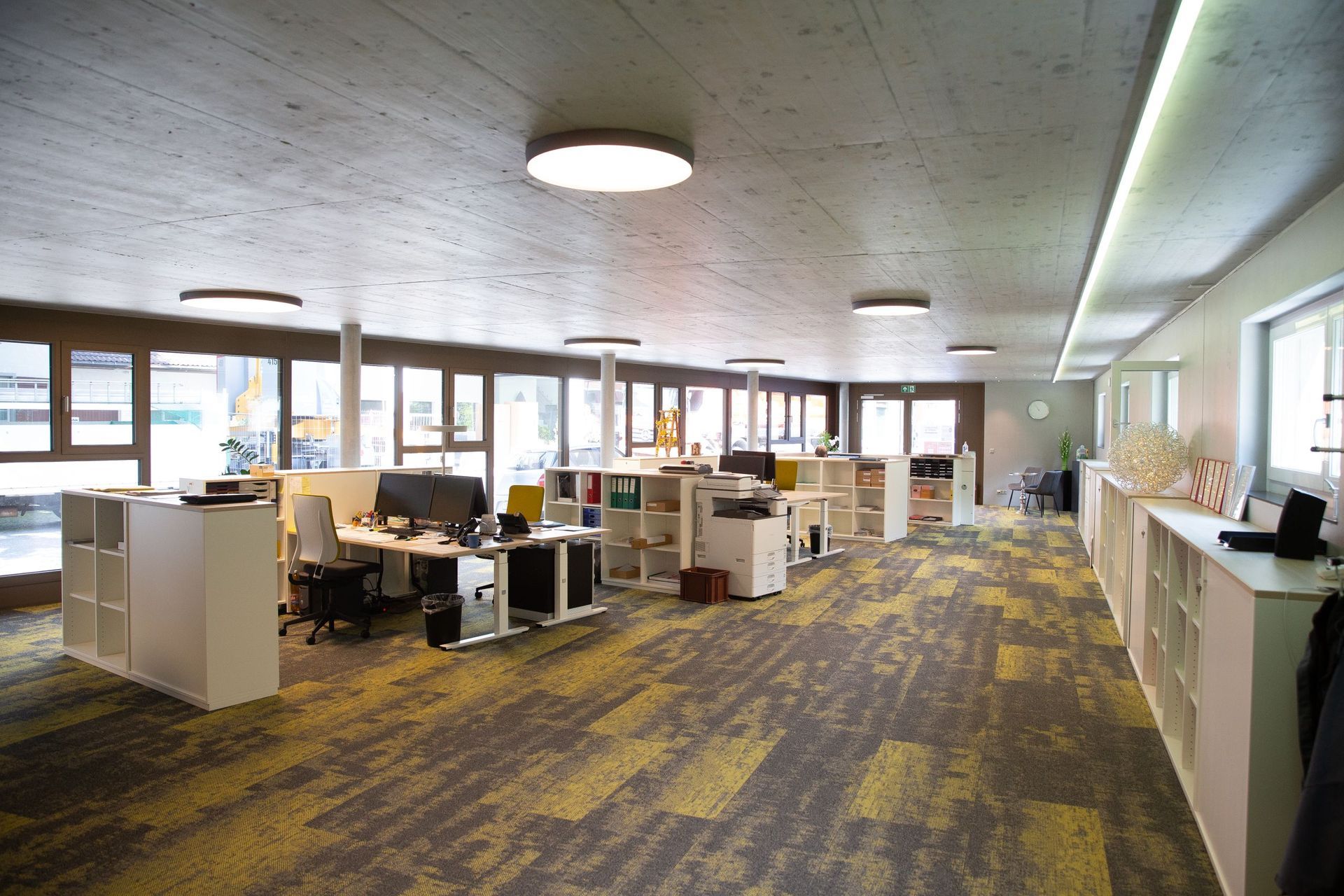 Heller Büroraum mit Arbeitsplätzen, Schreibtischen und Schränken. Lange Fenster, beige-gelb gemusterter Teppich und runde Dec
