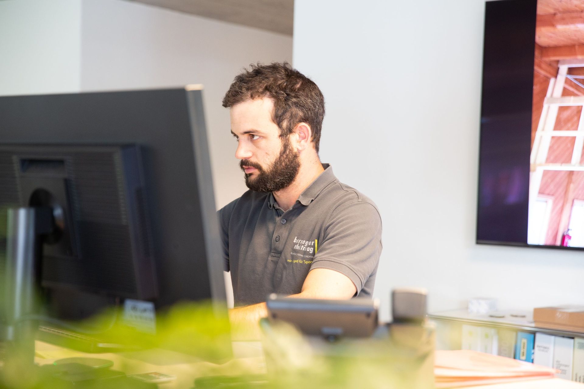 Mann mit Bart arbeitet an einem Computer an einem Büroschreibtisch. Er trägt ein graues Hemd und konzentriert sich auf 